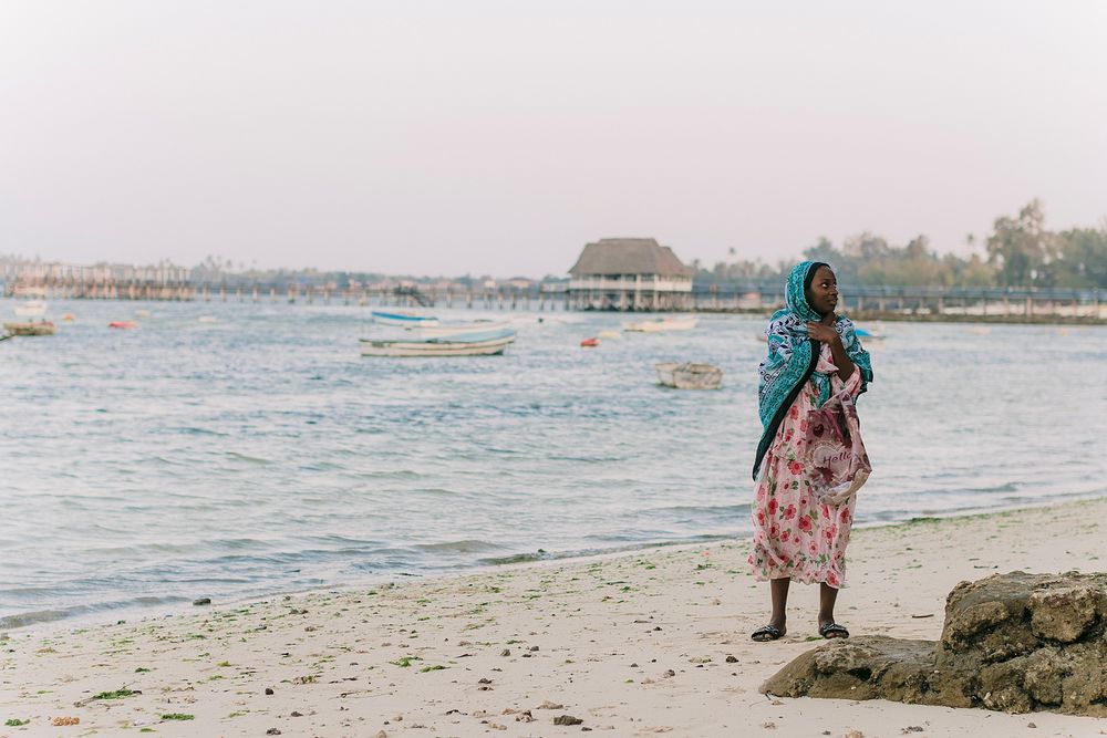 A woman in a floral dress and blue headscarf stands on a sandy beach with calm ocean waters behind her.