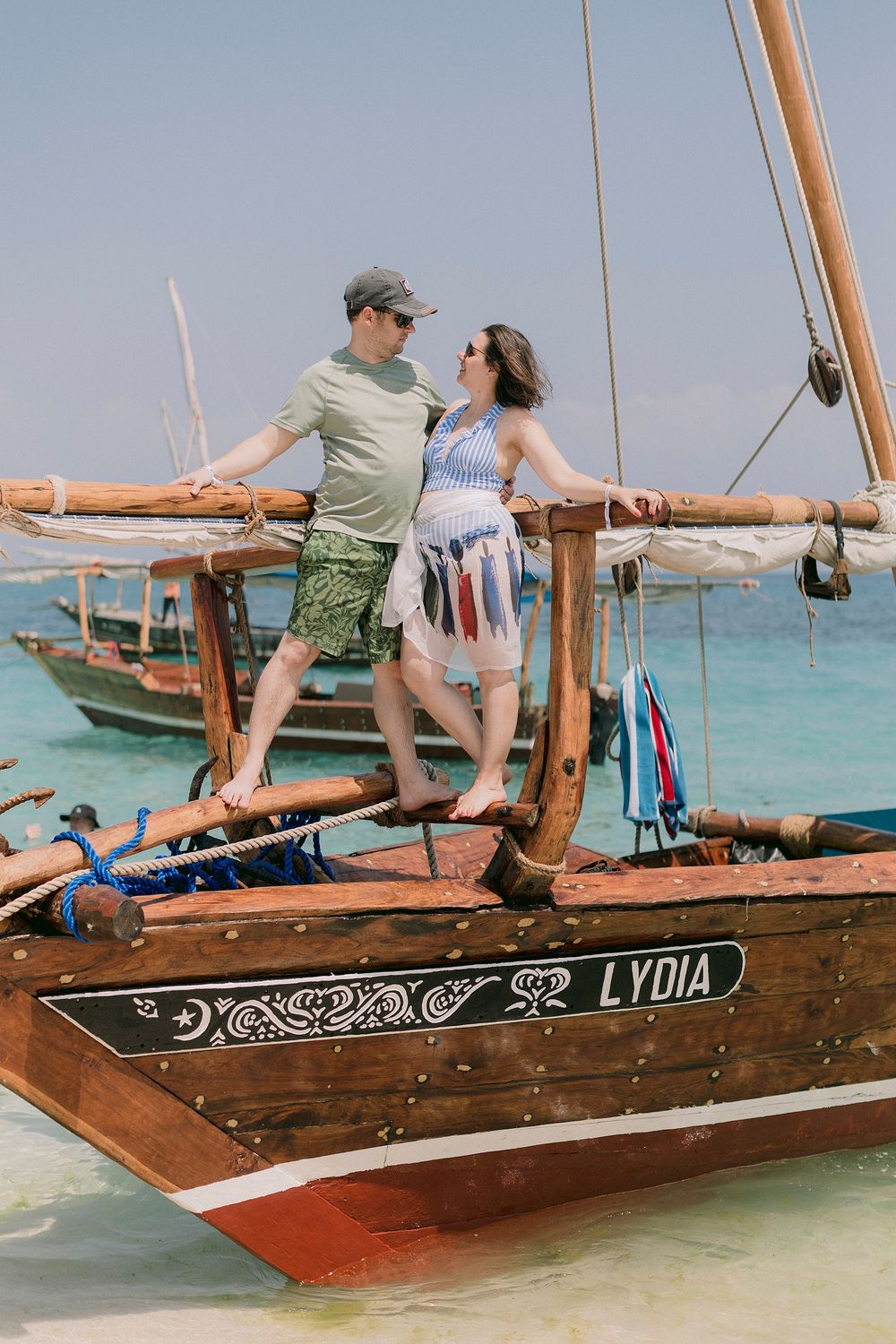 Couple standing on a wooden sailboat named Lydia in turquoise tropical waters on a sunny day.