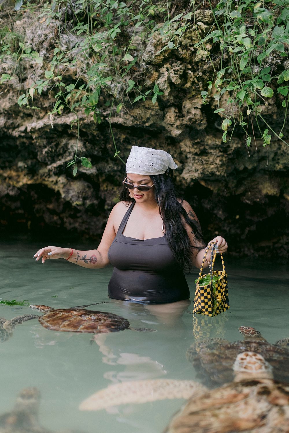 Woman in gray swimsuit and white bandana wading in turquoise water near sea turtles, holding beaded bag by rocky cliff.
