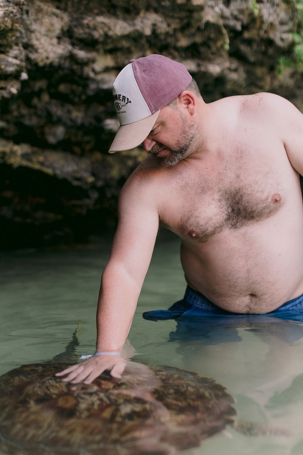 Shirtless man in a baseball cap touching a large rock or marine creature in shallow water near rocky cliffs.
