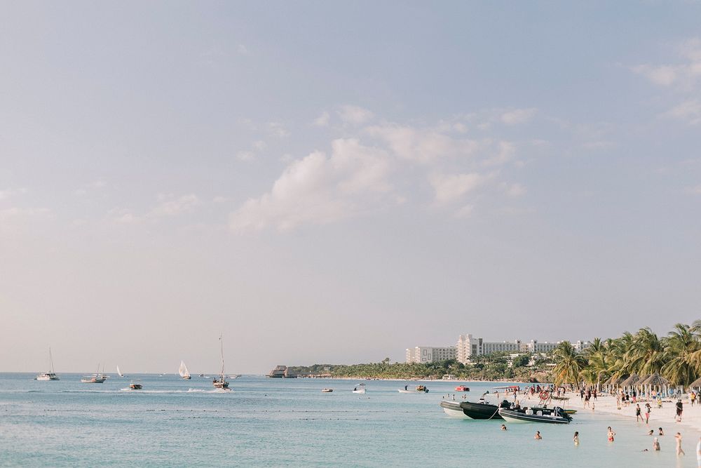 Tropical beach with calm turquoise water, boats, palm trees, and a city skyline under a partly cloudy sky.