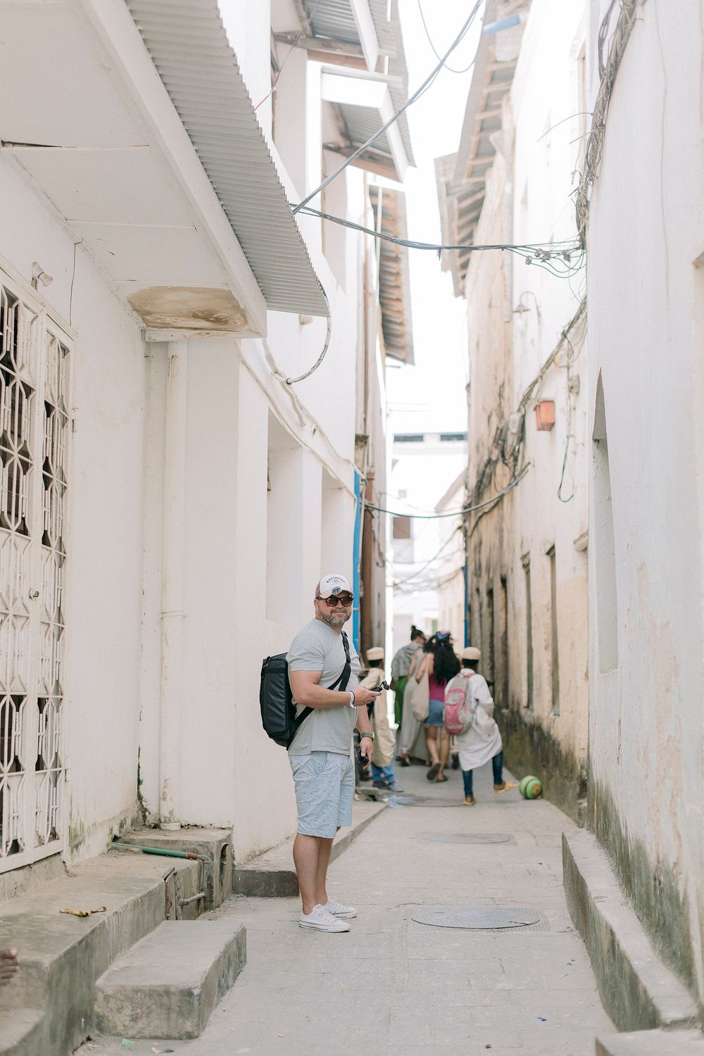 Tourist with backpack exploring a narrow white-walled alley in Stone Town, Zanzibar, with locals in the background.