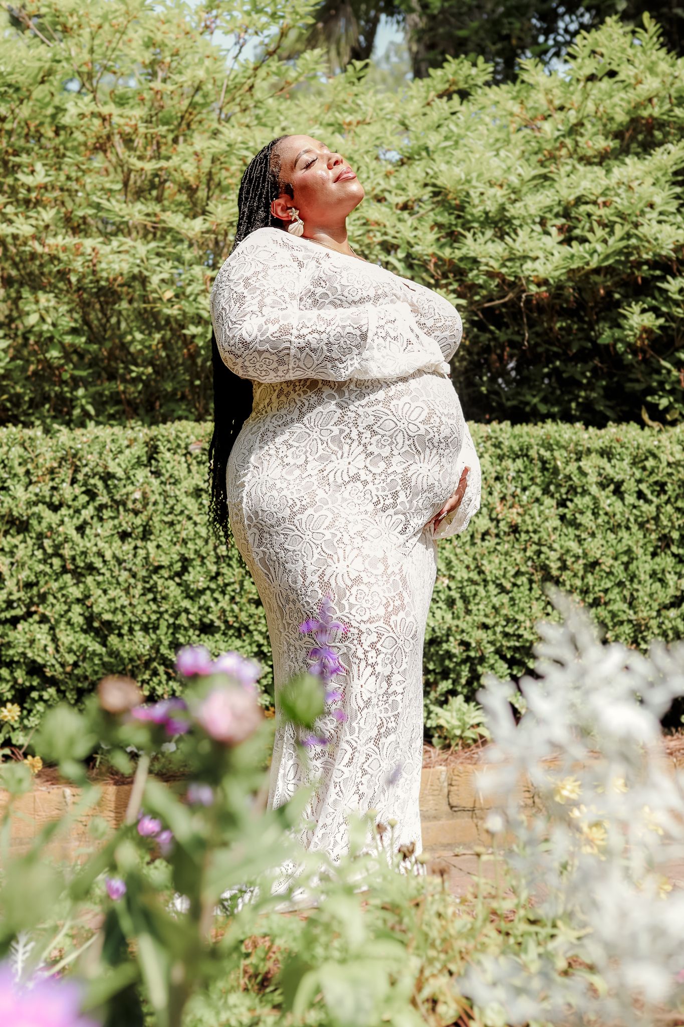 Young woman smiling in a sunlit garden, wearing a a white dress holding her pregnant belly, with tall pine trees in the background.
