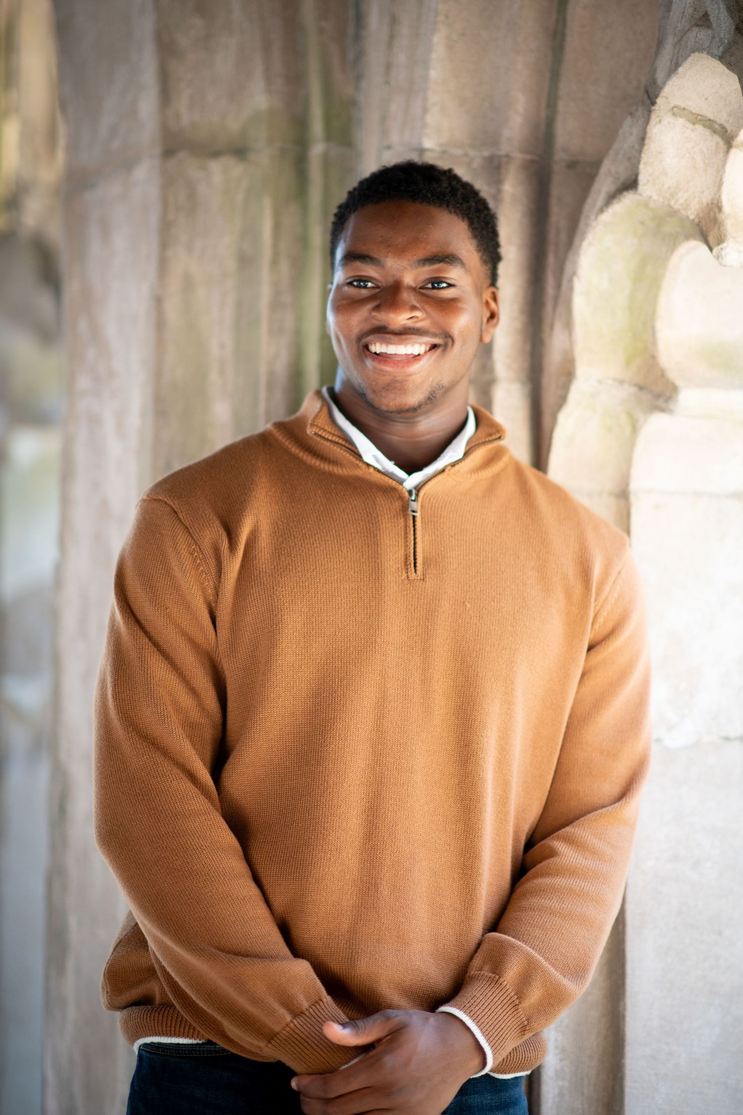 Smiling young man in a tan quarter-zip sweater standing outdoors near stone columns.