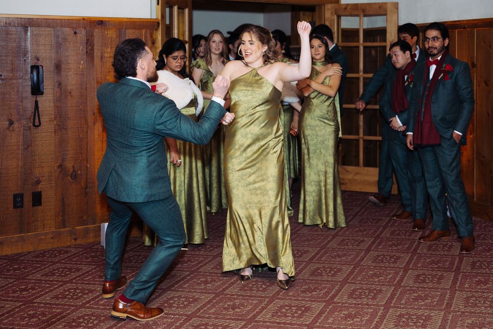 Wedding party dancing in green gowns and suits, celebrating joyfully in a wood-paneled reception hall.