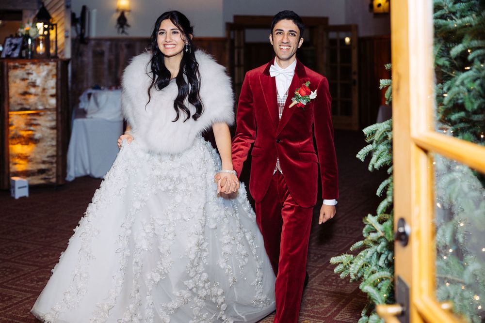 Bride in white floral gown with fur shawl holds hands with groom in red velvet suit at winter wedding reception.