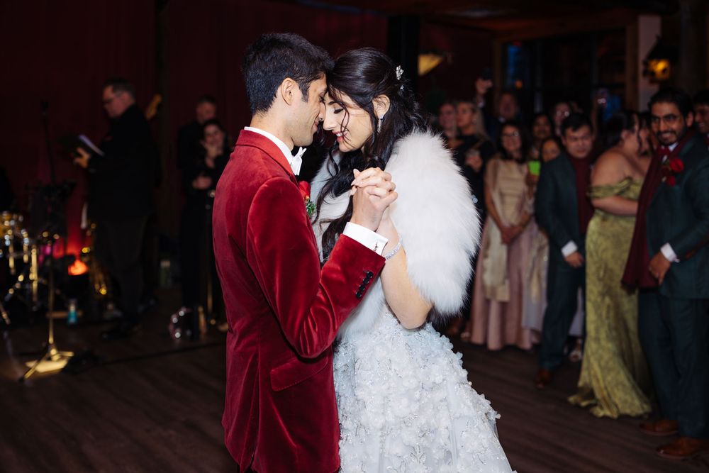 Couple shares first dance, groom in red velvet suit, bride in white feathered gown, guests watching in elegant venue.