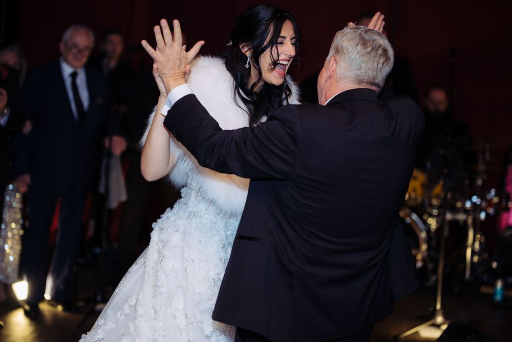 Bride in white ruffled gown dancing with older man in tuxedo at elegant wedding reception.