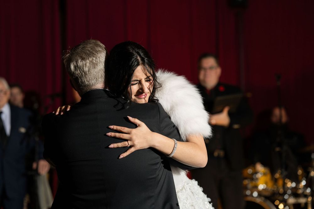 Couple embracing at elegant event, woman in white fur stole and black dress, red curtain backdrop.