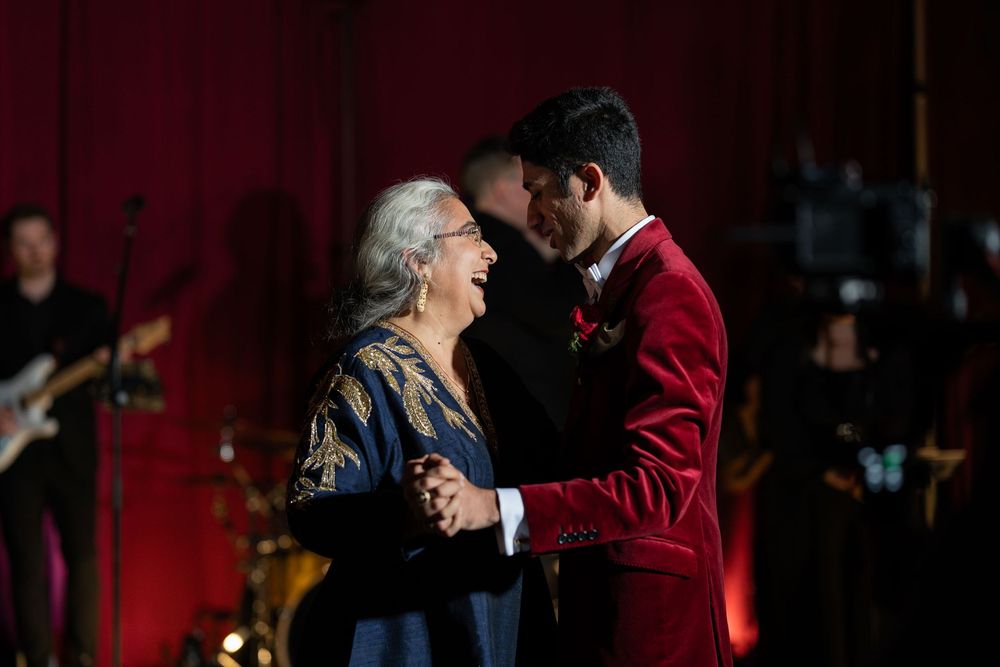 Groom in red velvet suit shares a tender dance with elderly woman at elegant wedding reception.