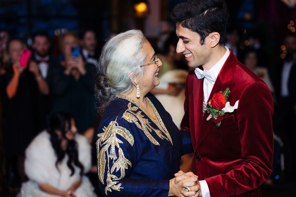 A young groom in red velvet tuxedo shares a joyful dance with an elderly woman at a wedding reception.