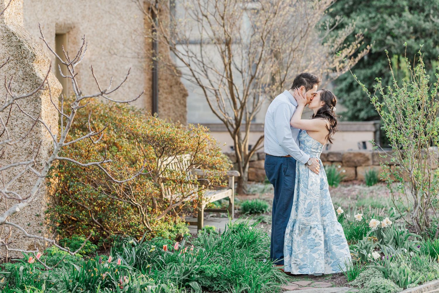 Couple sharing a romantic kiss in a lush garden setting, with the woman in a floral blue dress and man in navy suit.