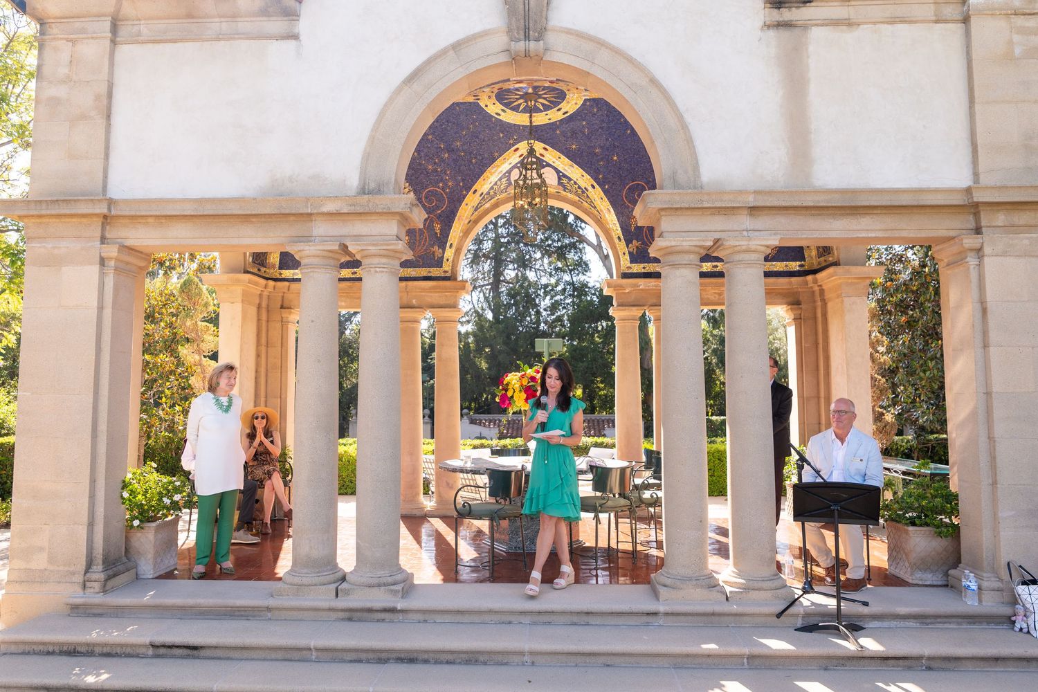 Woman in teal dress speaks at elegant columned pavilion ceremony with ornate arch and garden backdrop.