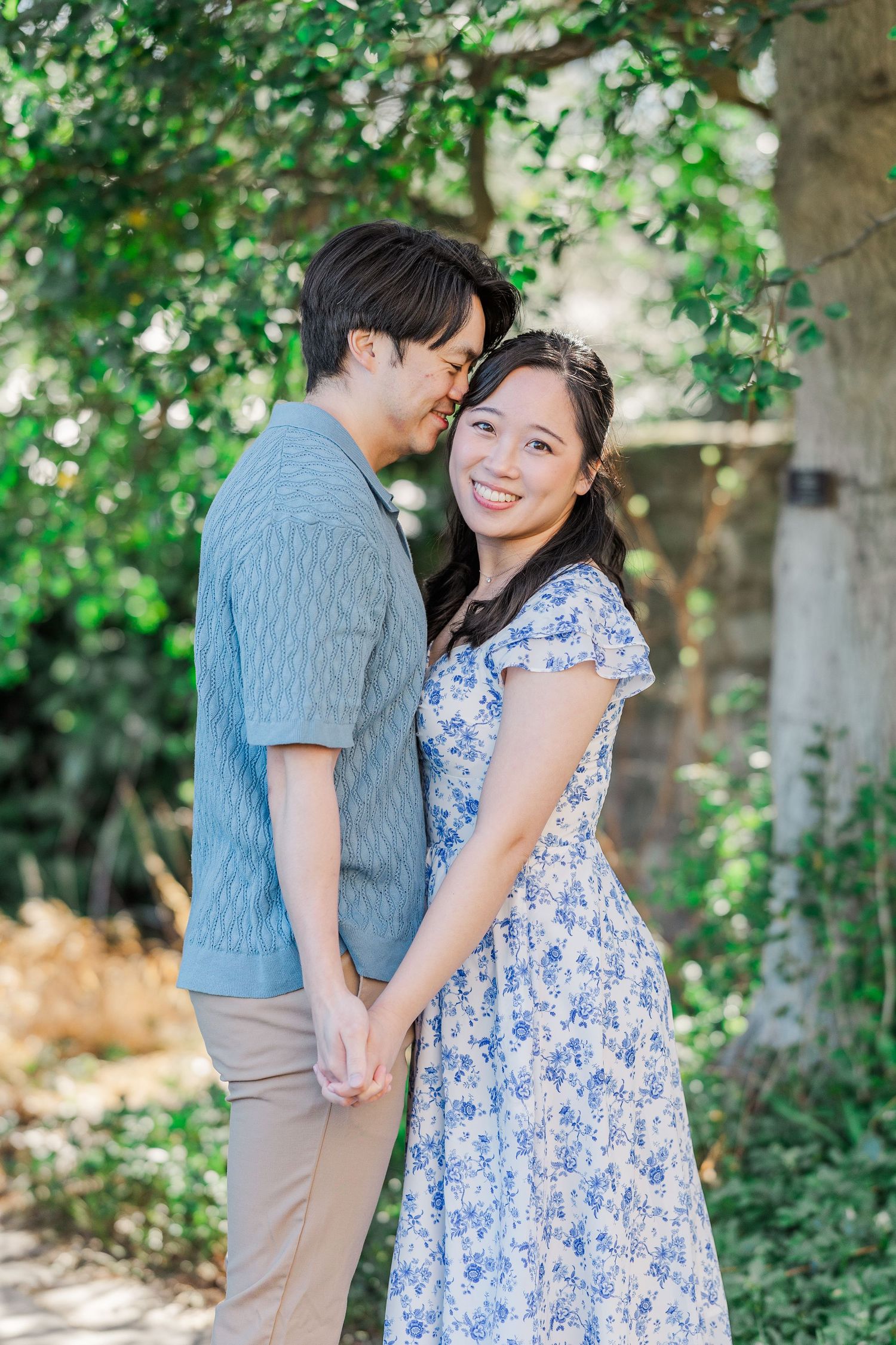 A smiling couple holds hands in a lush garden, woman in floral dress, man in blue shirt, sharing a tender moment.