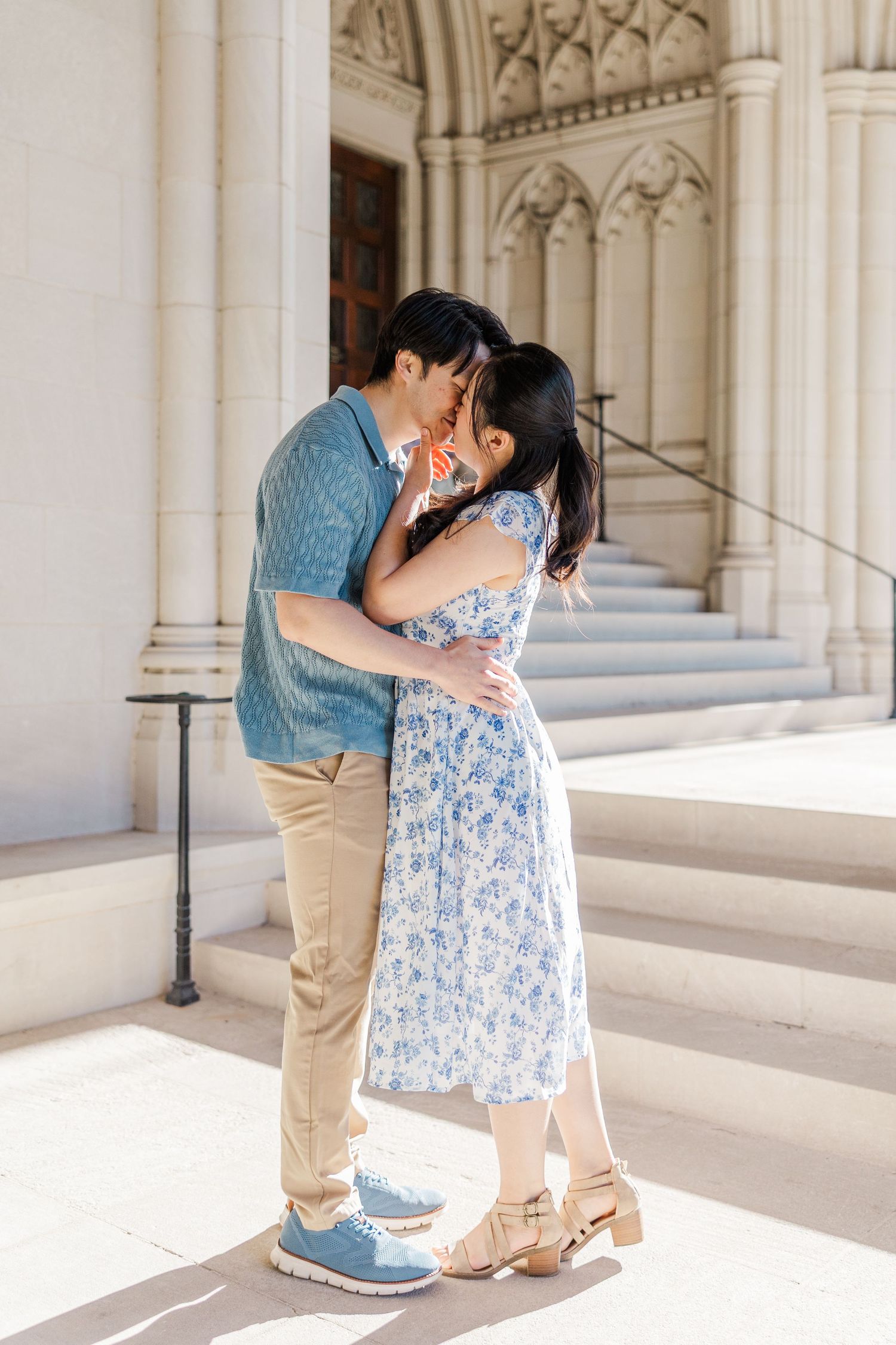 Couple sharing a romantic kiss in front of a grand stone building with ornate Gothic architecture on a sunny day.
