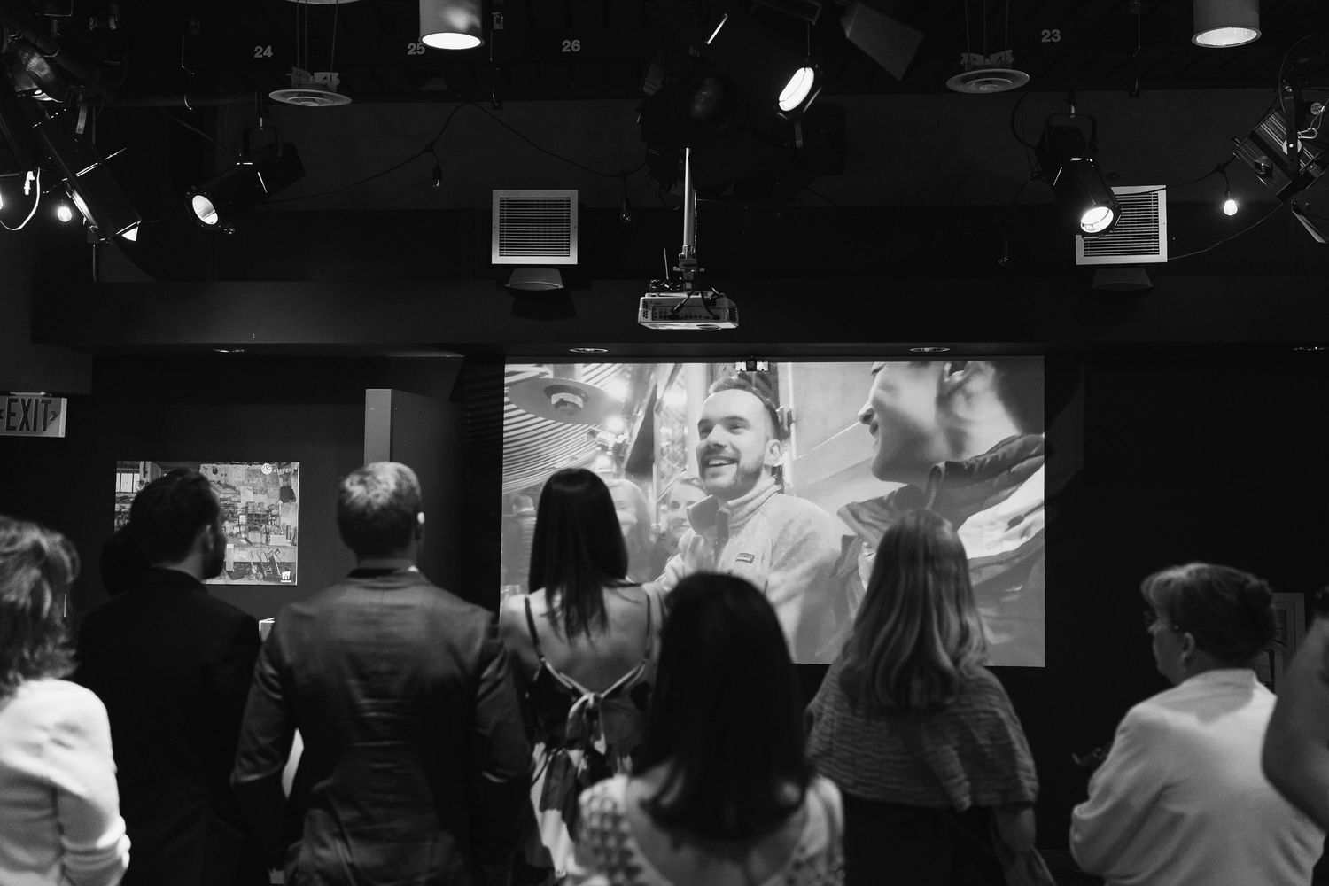Black and white photo of audience members watching a presentation screen displaying a smiling man at an event.