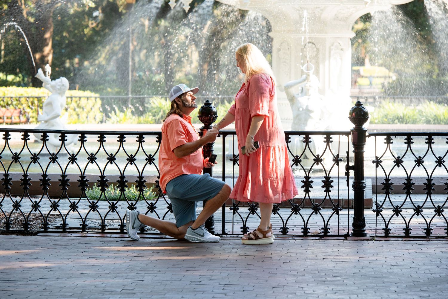 Man kneeling on one knee proposing to woman in pink dress near ornate fountain in sunny park setting.
