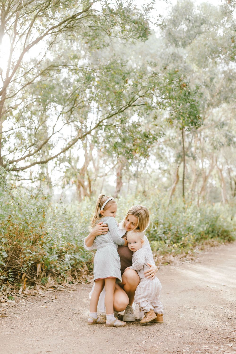 A mother crouches on a dirt path hugging her two young daughters in a sunlit outdoor setting.
