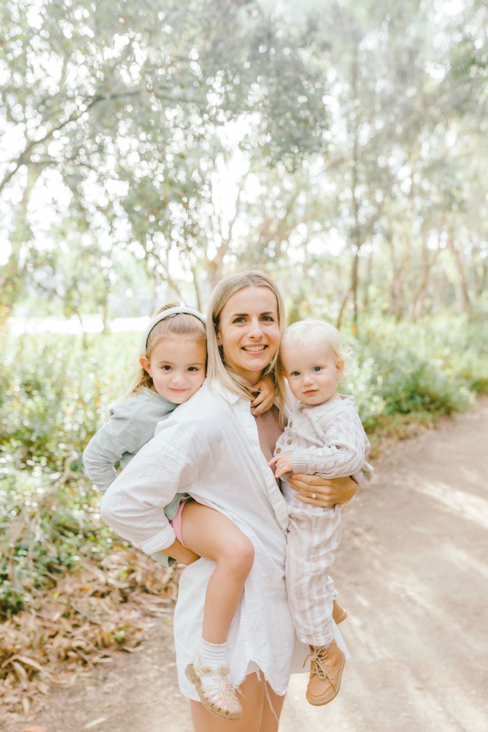 A smiling blonde mother holds two young children on a sunny dirt path surrounded by lush green trees.