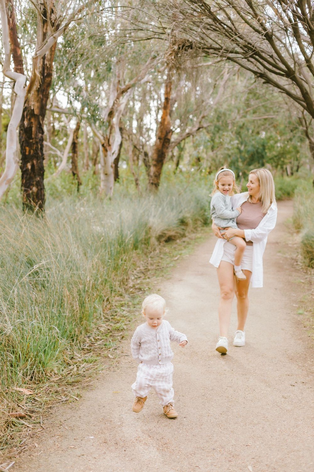 A mother carries a toddler while walking a scenic bush path as a young child toddles ahead in natural light.