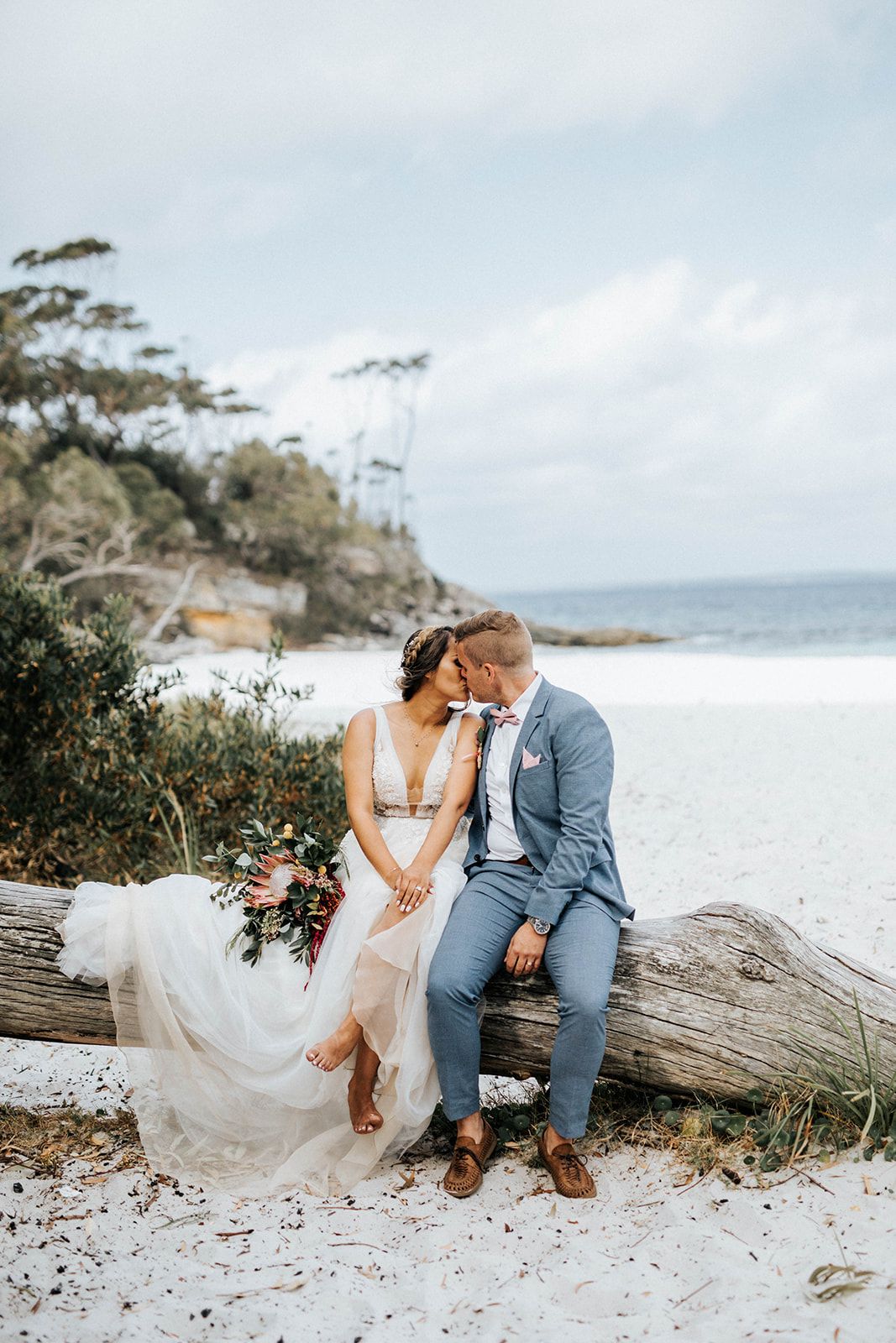 Bride and groom share a romantic moment sitting on driftwood at a white sand beach, bride in flowing gown holding bouquet.