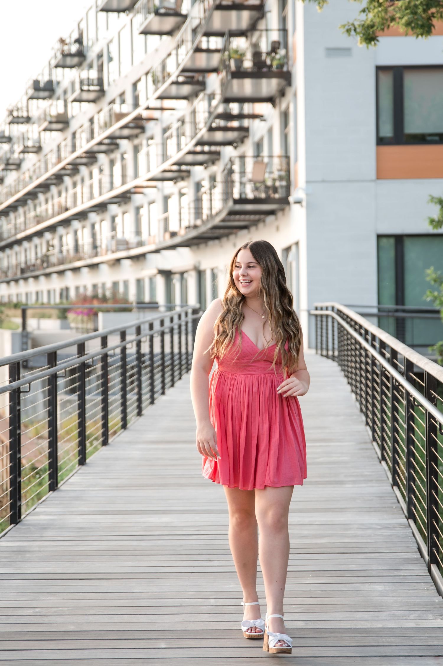 Young woman in pink dress smiling on wooden walkway beside modern apartment building on sunny day.