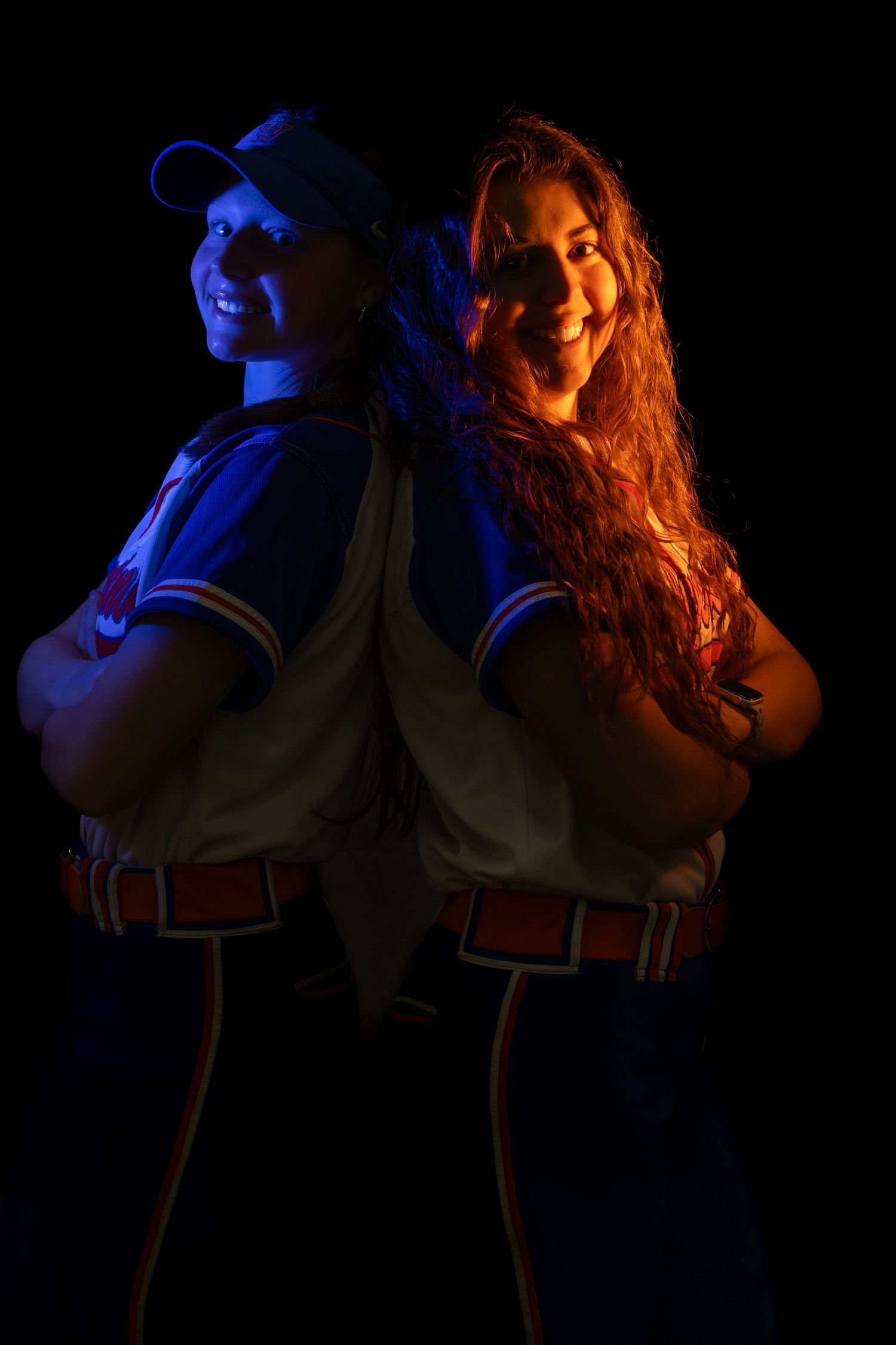 Two softball players in uniforms pose back-to-back, lit dramatically in blue and orange light against a dark background.
