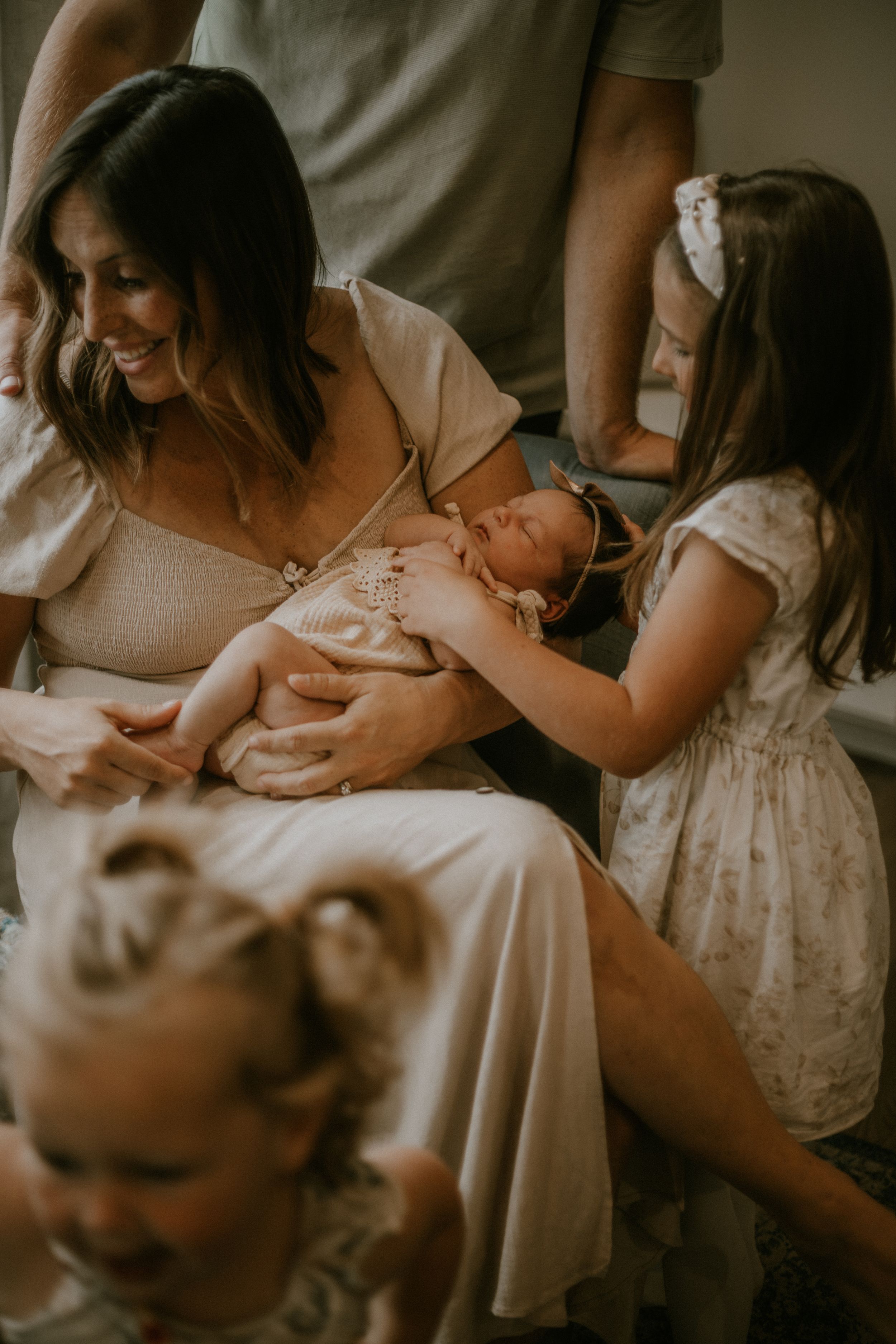 A mother smiles while holding her newborn baby, surrounded by young daughters in a warm, candid family moment.