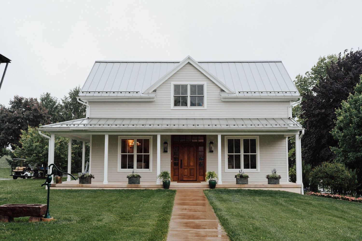 White farmhouse with metal roof, covered porch, wood door, and lush green lawn on a cloudy day.