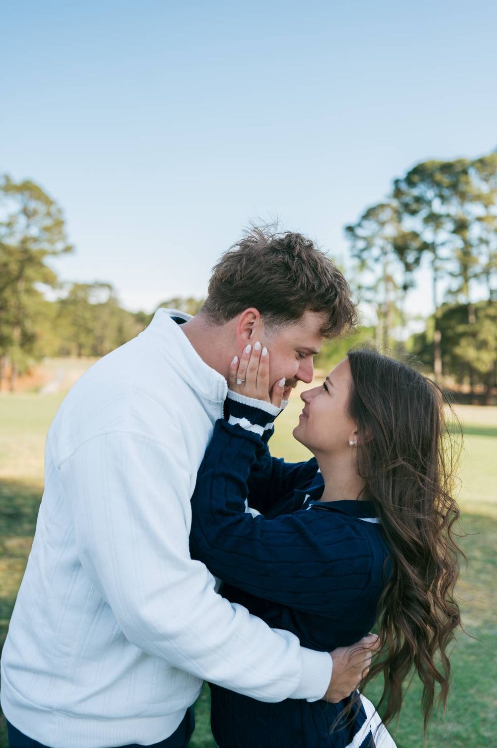 Couple sharing a tender almost-kiss embrace outdoors on a sunny golf course with trees behind, lifestyle photographer High Point NC