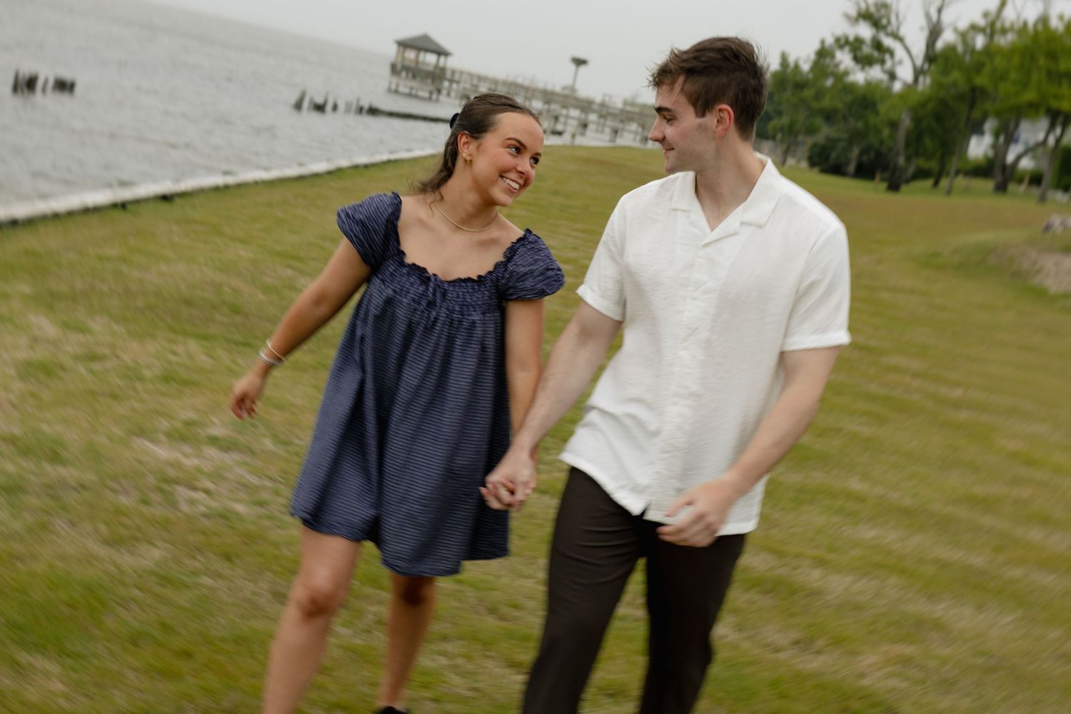 Black and white photo of a laughing couple holding hands and running barefoot on a beach, woman in white dress.