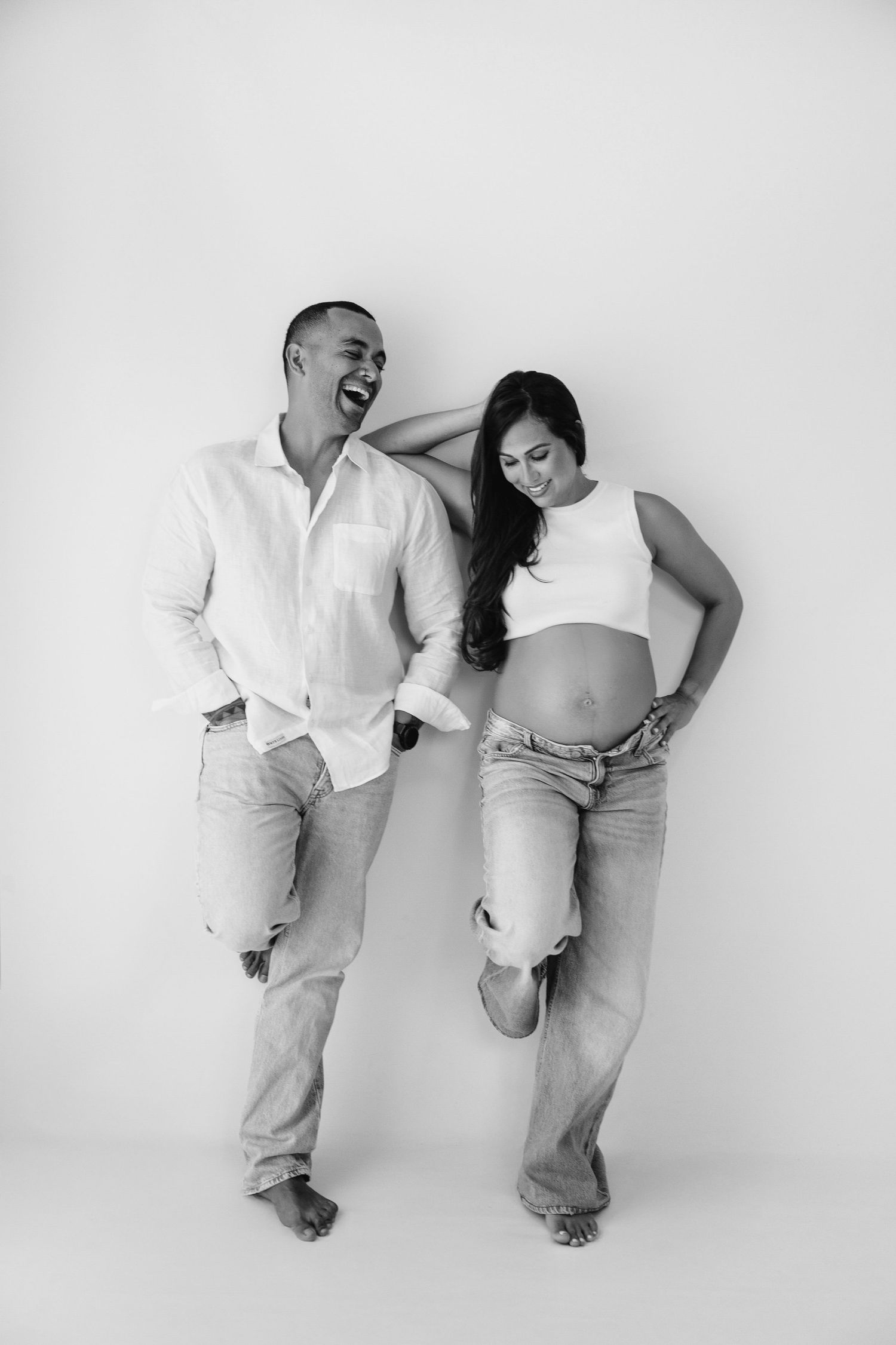 Smiling pregnant couple in black tops with dog on white bed, man with tattoo touching woman's shoulder.