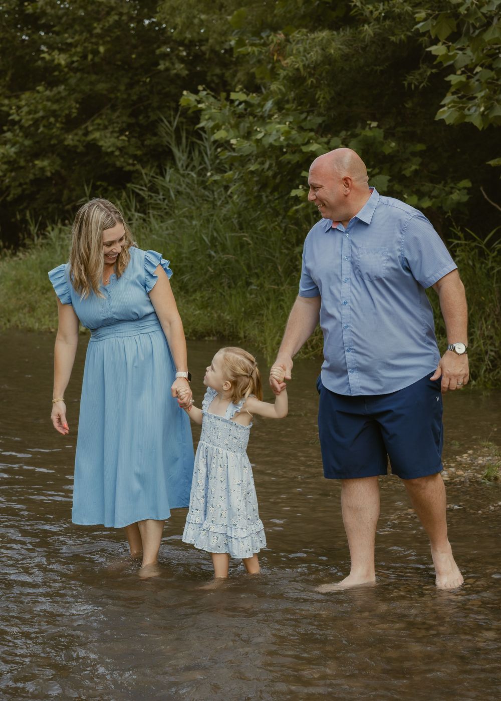 A couple in blue outfits holds hands with their young daughter while wading in a shallow creek outdoors.