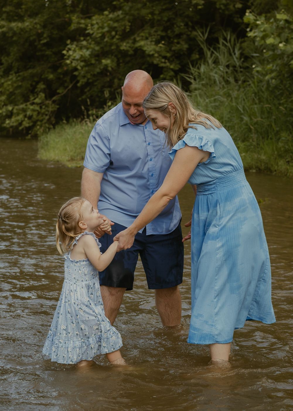 Parents in blue outfits smile at their toddler daughter while wading together in a shallow creek.