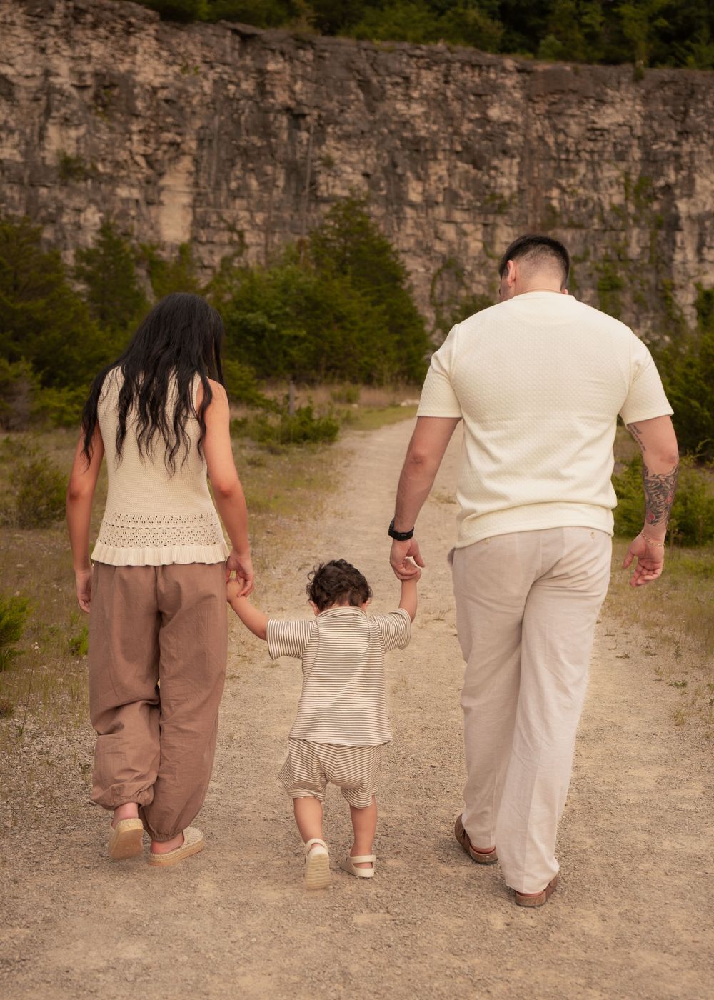 Family of three walking hand-in-hand on a dirt path near a stone wall, parents holding toddler's hands.