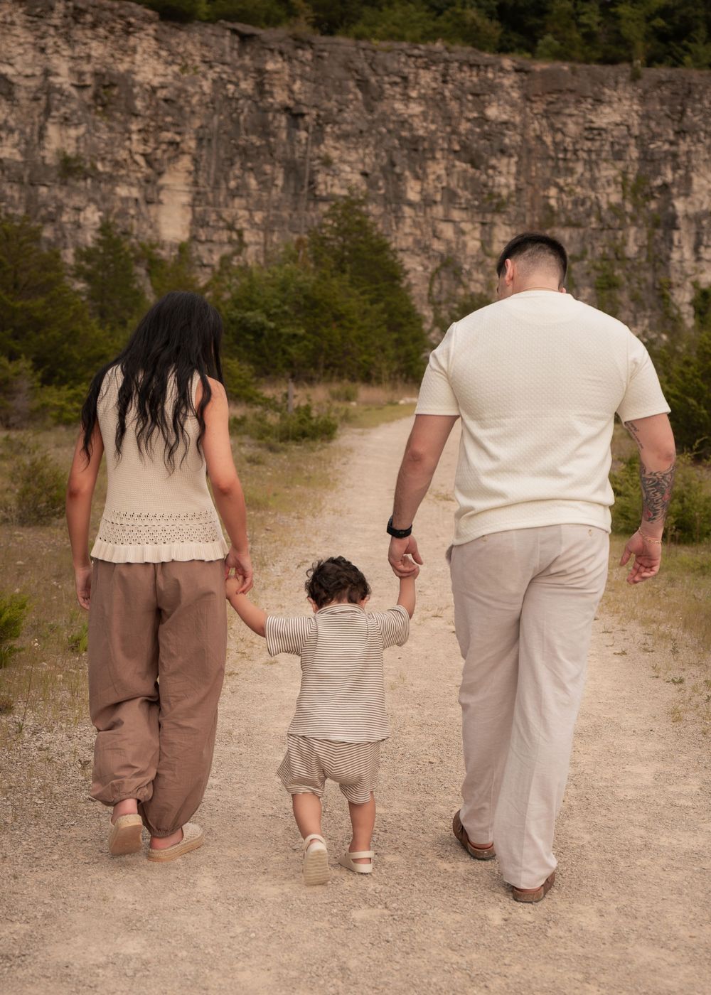 Family of three walking hand-in-hand along rocky outdoor path, seen from behind.