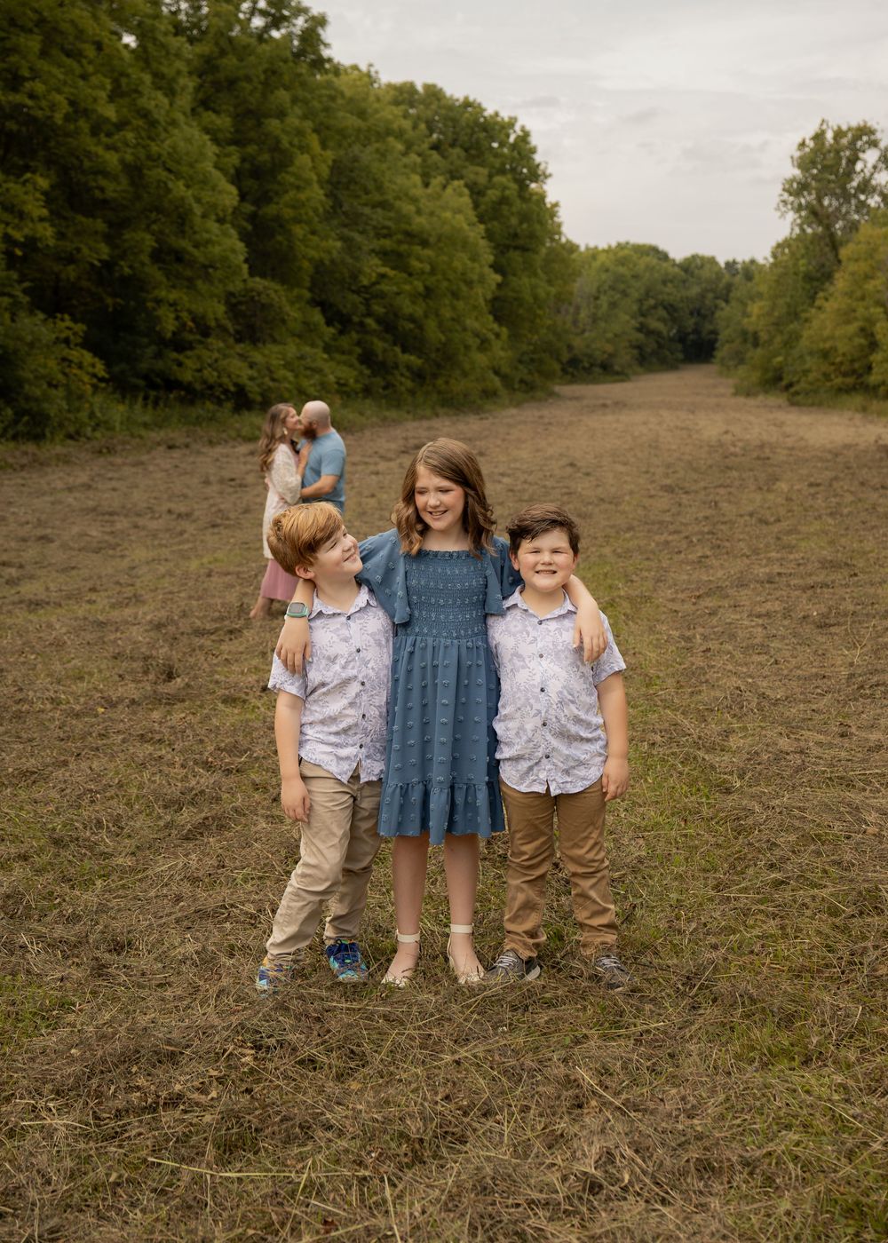 Three children pose in an open field, with two adults embracing in the background surrounded by lush green trees.