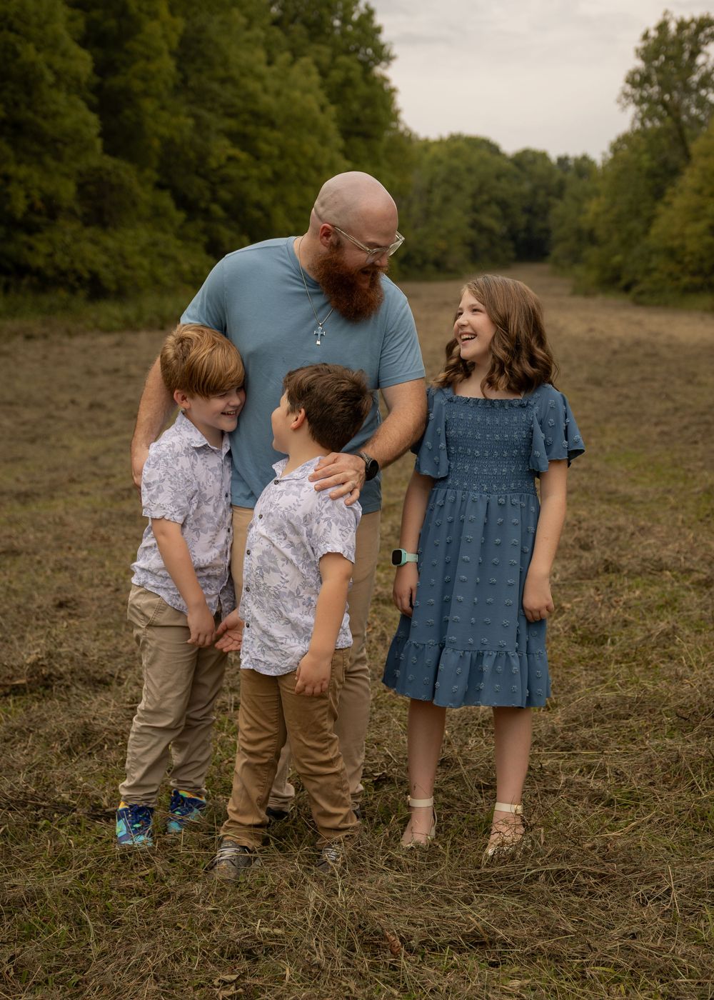 A bald bearded father laughs with his three young children in a grassy field surrounded by trees.