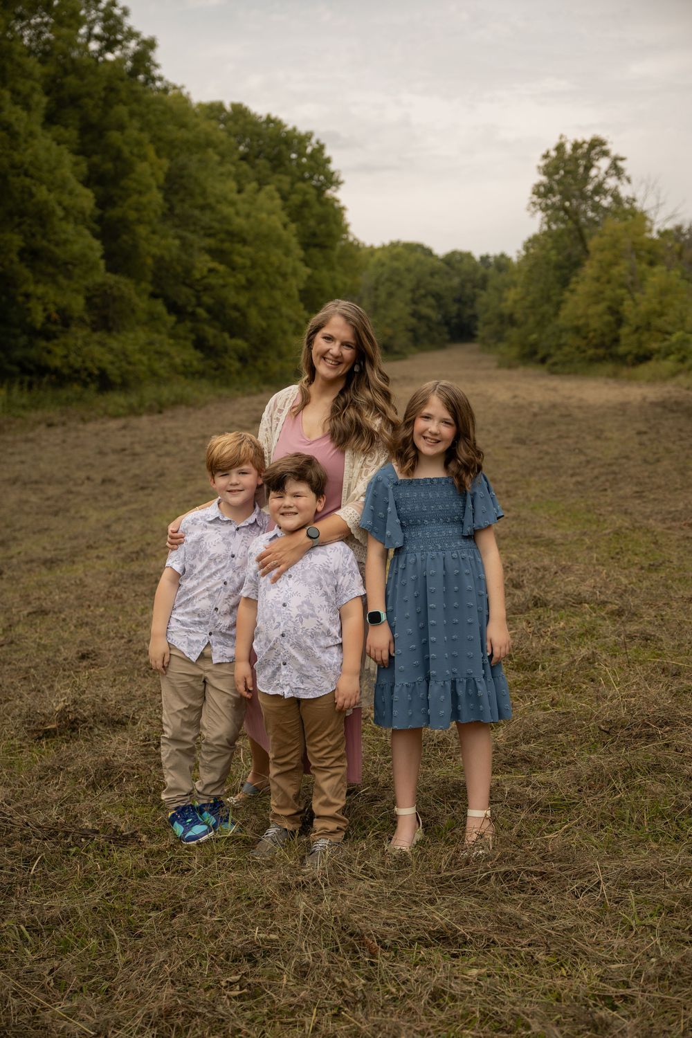 A woman and three children pose together in an open field surrounded by lush green trees during an outdoor family photo session.