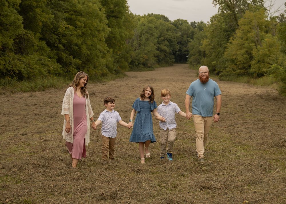 Family of five walking hand-in-hand through a scenic meadow surrounded by lush green trees during an outdoor photo session.