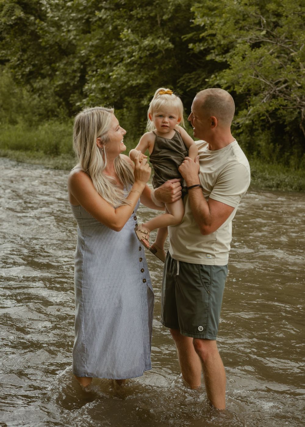 Young family of three wading in a shallow creek, father holding toddler while mother smiles outdoors.