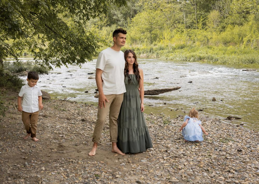 A tall couple stands barefoot on a rocky riverbank while two young children play nearby in nature.