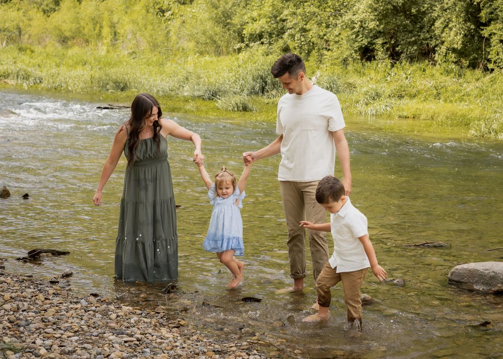 A family of four holds hands and splashes through a shallow rocky creek on a sunny summer day.