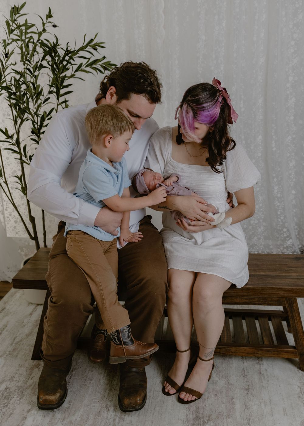 Family of four with newborn baby sitting on wooden bench in bright studio setting.