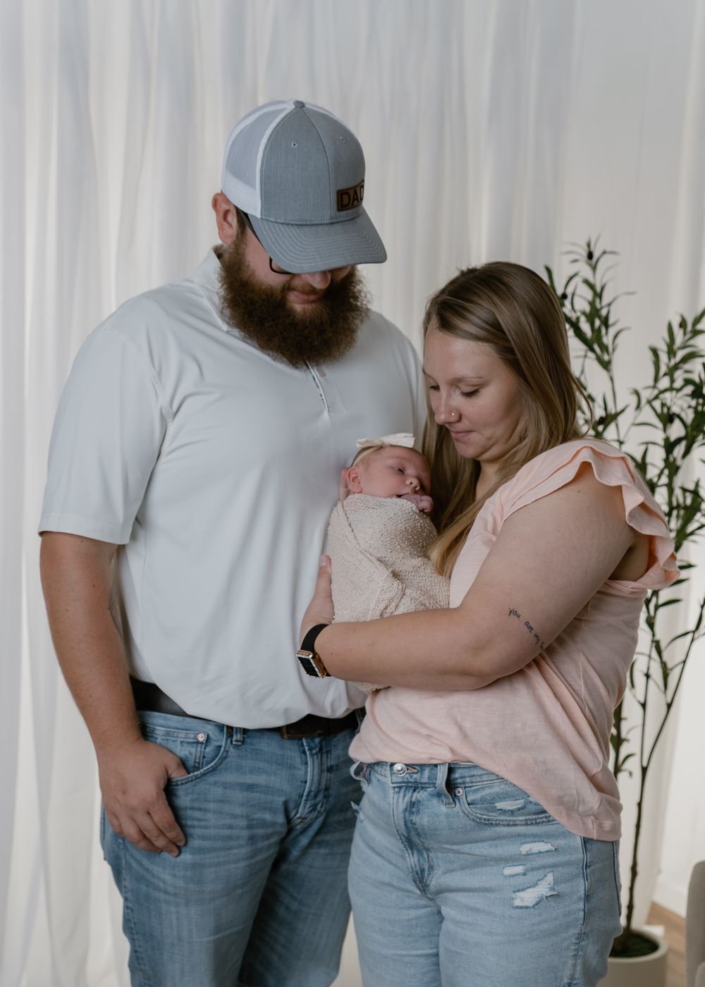 New parents in casual attire gazing lovingly at their newborn in white studio.