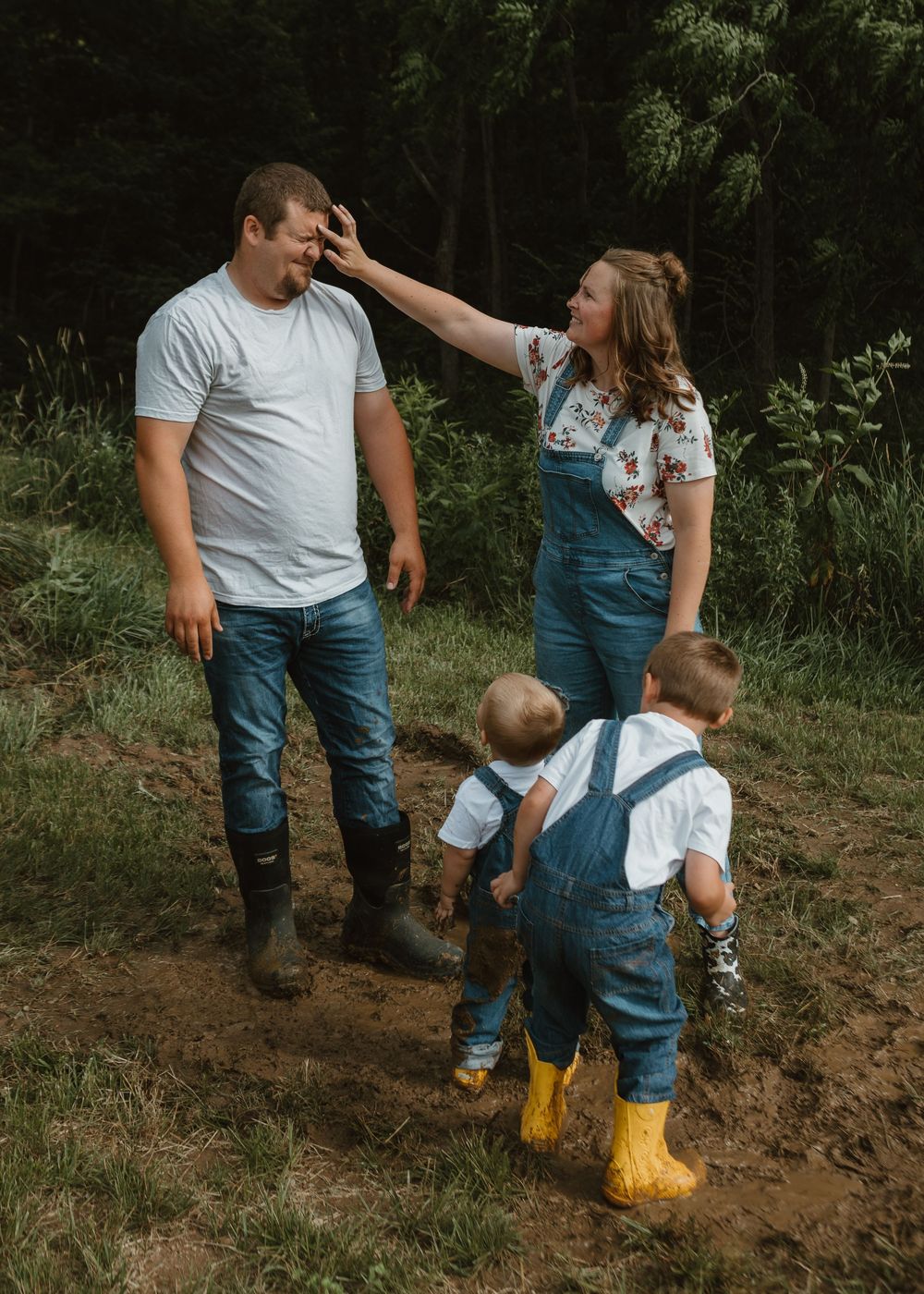A young couple in denim overalls and two toddlers in yellow boots play together in a muddy outdoor setting.