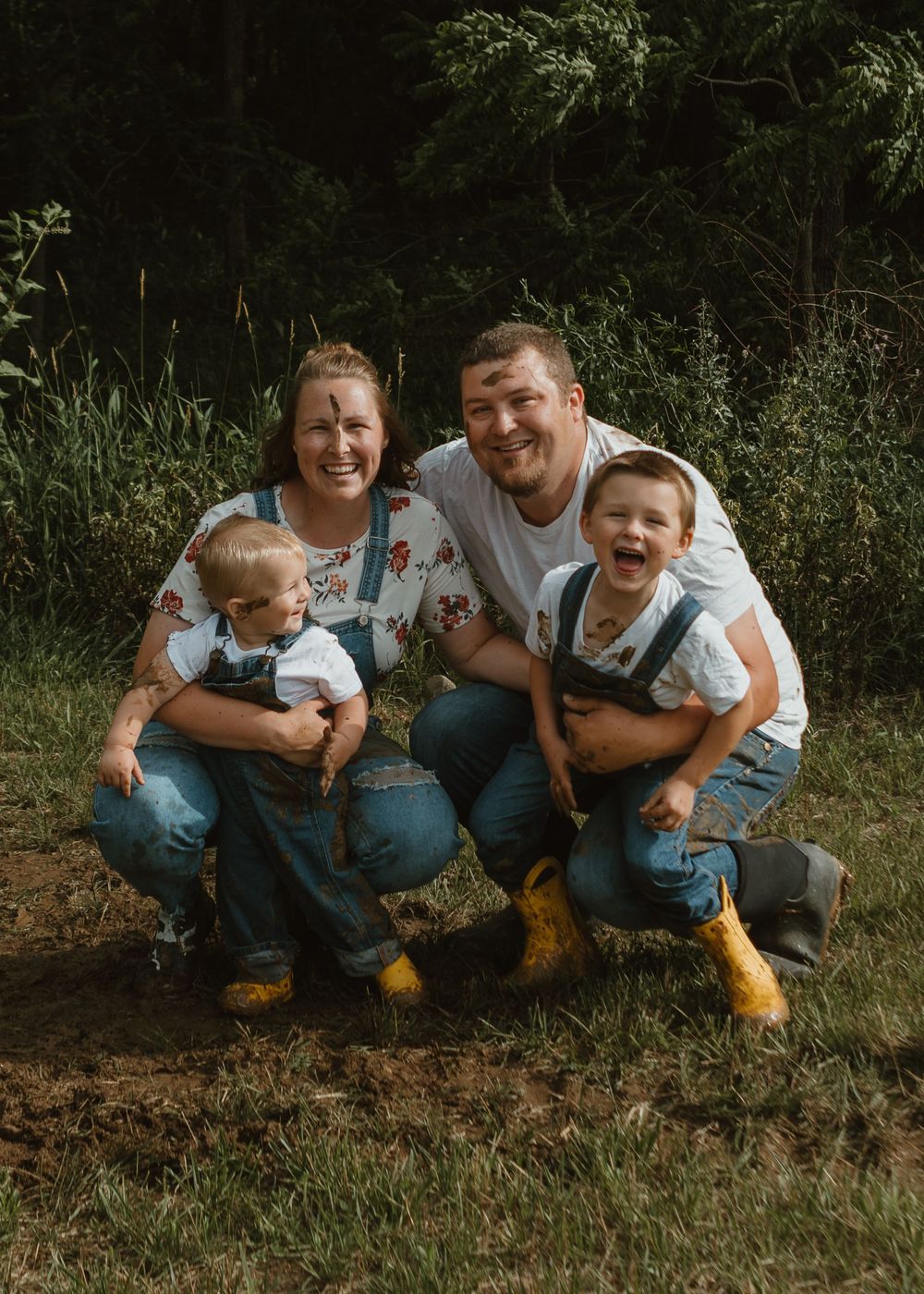 A cheerful family of four in matching denim overalls and yellow boots poses together outdoors in the grass.