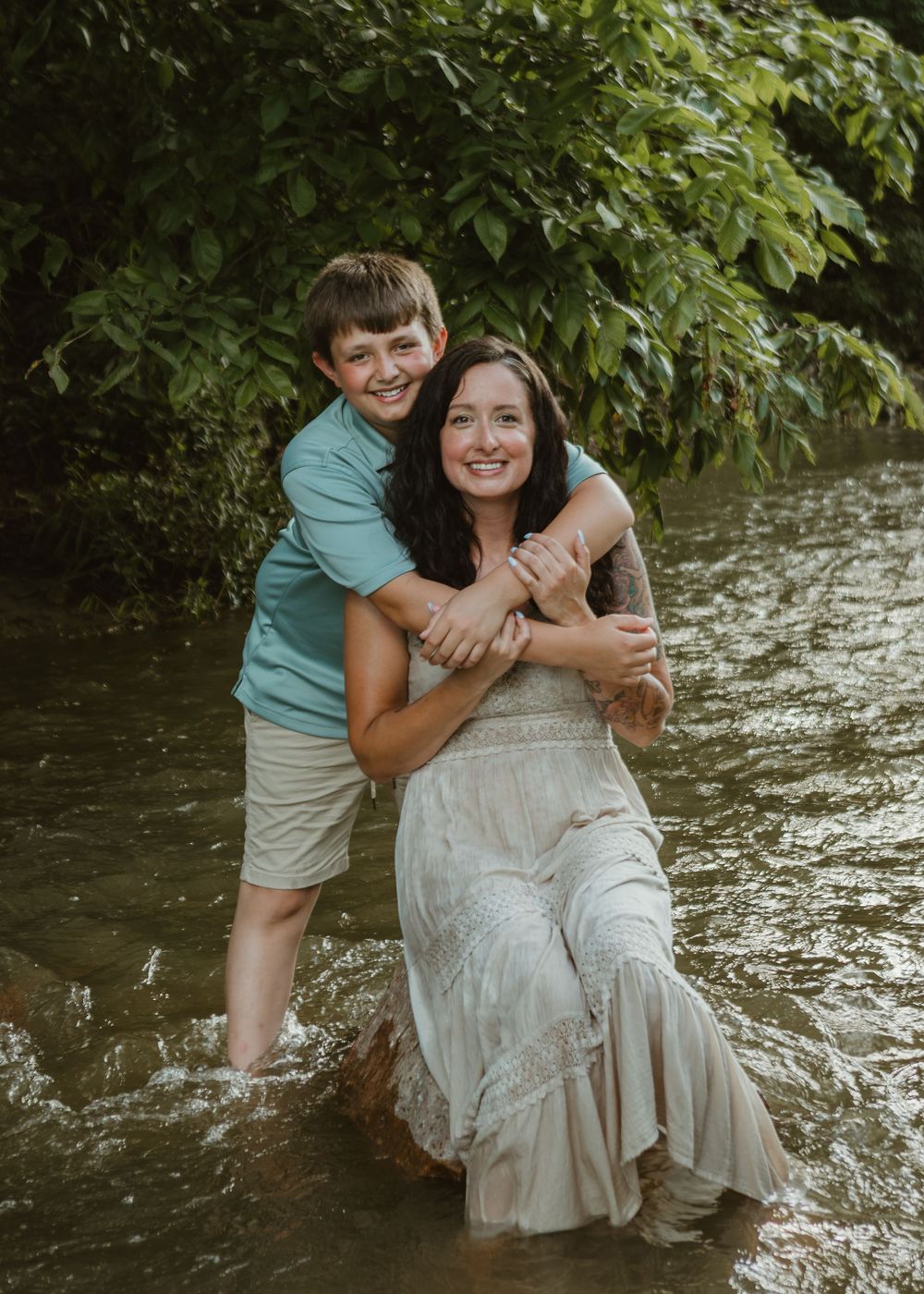 A smiling boy hugs his mother from behind as they stand together in a shallow creek surrounded by greenery.
