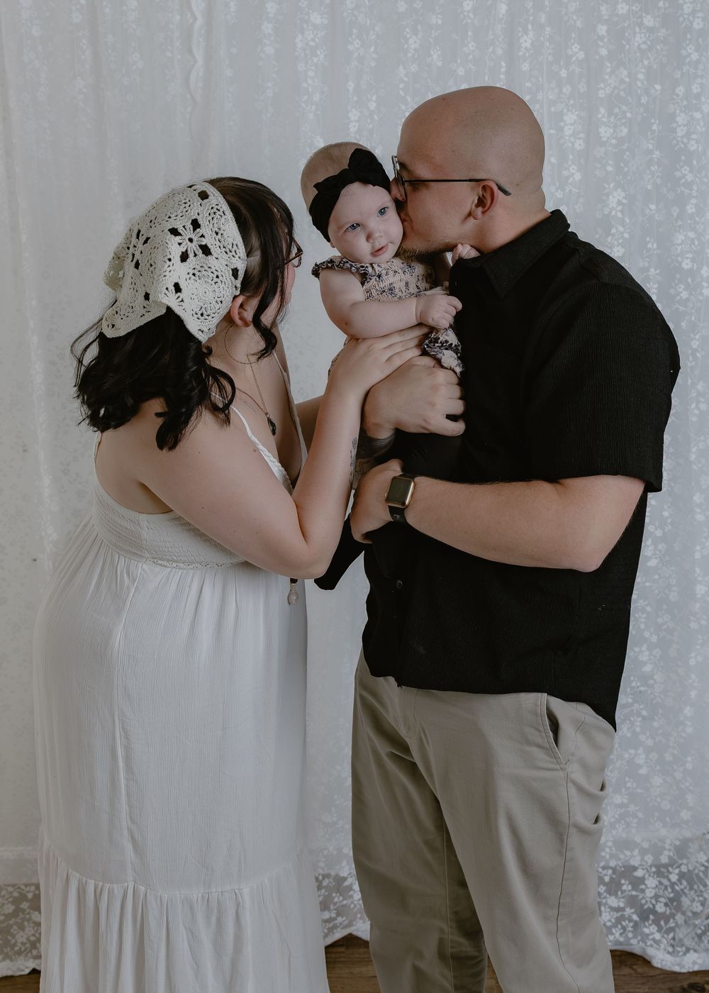 Parents in black and white outfits kissing their baby girl in bright studio.