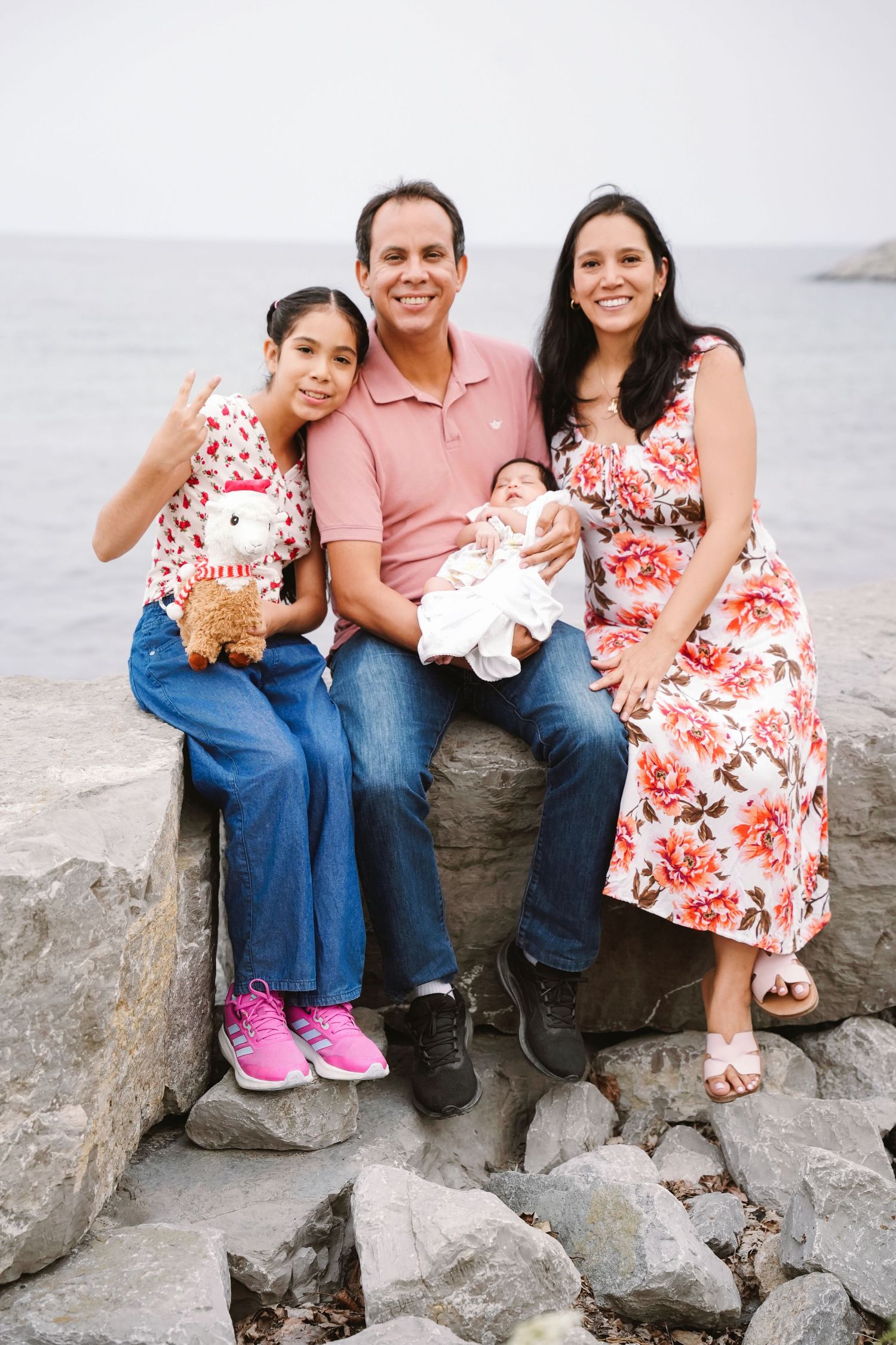 Happy family of four posing on rocky shoreline, parents with young daughter and newborn baby by the ocean. Toronto GTA